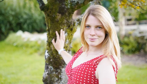 Young woman with blonde hair wearing a red polka dot dress, leaning against a moss-covered tree in a garden setting.