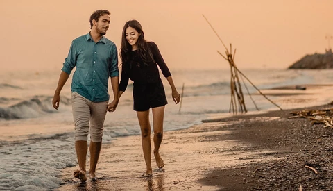 Couple walking barefoot along the shoreline at sunset, holding hands on a beach.