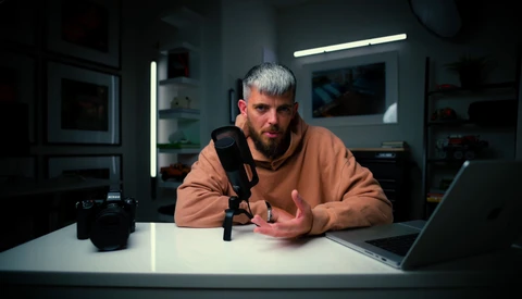 Man speaking into microphone at desk with laptop in dimly lit studio setup.