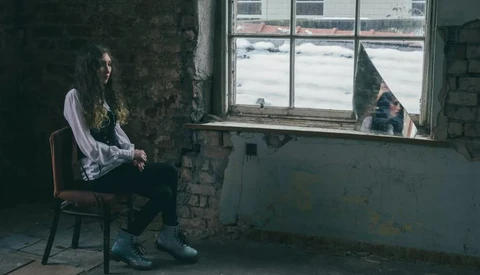 Person with long dark hair sitting on a wooden chair in a deteriorating room, gazing toward a large window overlooking water.