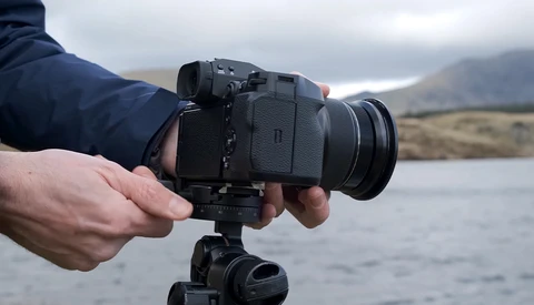 Photographer holding a mirrorless camera on a tripod overlooking a mountain lake.