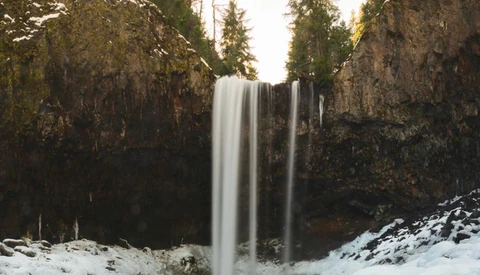 Waterfall cascading between tall rock cliffs surrounded by evergreen forest and snow.
