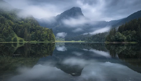 Mountain lake with forested shores and misty peaks reflected in calm water.