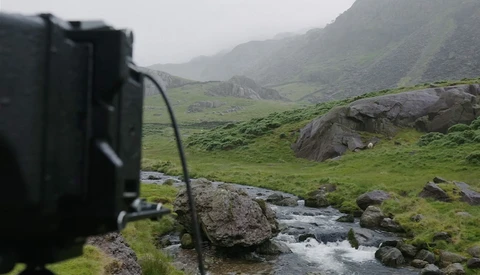 Camera mounted on tripod overlooking a misty mountain valley with a stream and green meadows.