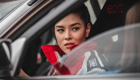 Woman in red jacket sitting in car's driver seat, looking toward camera through windshield.