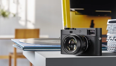 Leica M rangefinder camera with 50mm lens on a white desk with blue notebook and yellow wall backdrop.