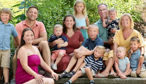 Multi-generational family group of 15 people posed together outdoors against green foliage.