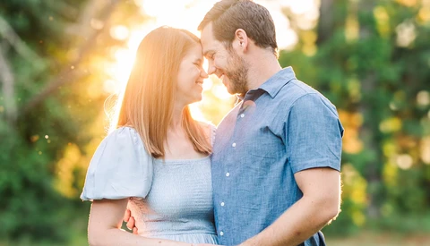 Couple embracing outdoors with foreheads touching, backlit by golden sunlight filtering through trees.