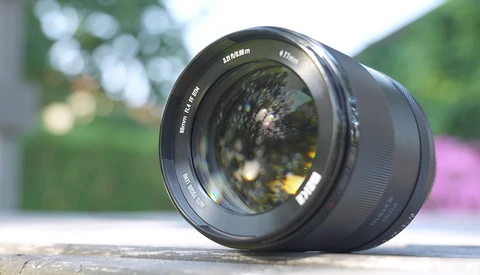 Black prime lens positioned on wooden surface with blurred garden and flowering plants in background.