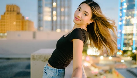 Woman in black crop top and jeans posing on rooftop at dusk with city skyline behind her.