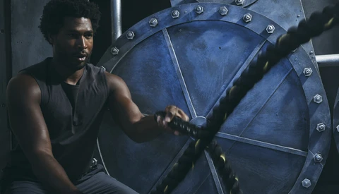 Man in dark tank top sits against large blue industrial gear wheel with metal studs and rivets.