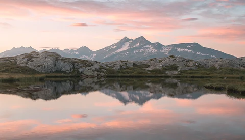 Snow-capped mountain reflected in still lake water at sunset with pink and orange sky.