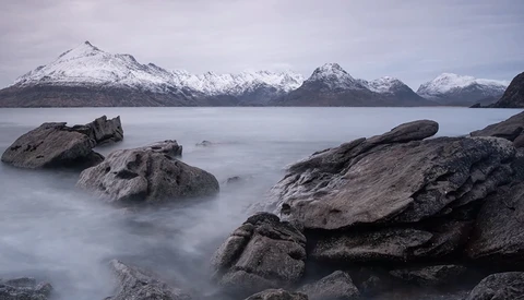 Snow-capped mountains across a calm fjord with foreground rocks smoothed by long-exposure water effect.