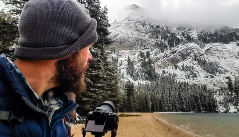Bearded photographer in winter beanie holding camera equipment overlooking snow-covered alpine lake and mountains.