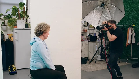 Photographer adjusting camera settings while shooting a portrait of a person seated against a white wall in a studio setting.
