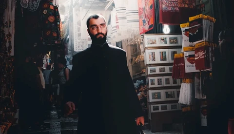 Bearded man in dark coat walking through narrow marketplace corridor lined with merchandise displays.