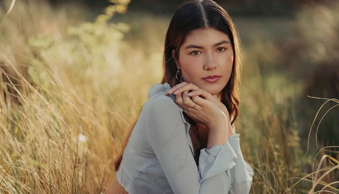 Young woman in light blue shirt posing in golden grass field during golden hour.