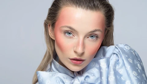 Portrait of a woman with blue eyes and pink-toned cheeks against a neutral gray background.