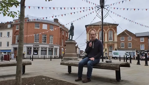 Person sitting on a wooden bench in a town square surrounded by Victorian-era brick buildings and festive bunting.
