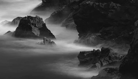 Rocky coastal formations emerging from mist and turbulent seawater in monochrome.