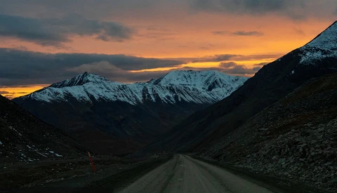 Straight gravel road stretching toward snow-capped mountains under a golden sunset sky.