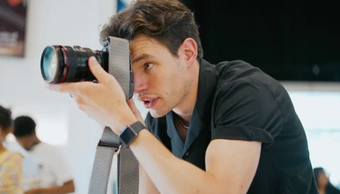 Photographer holding a professional camera with telephoto lens up to his eye while composing a shot indoors.