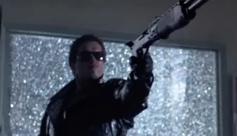Person in sunglasses and dark clothing aiming a rifle with scope toward bright, sparkly windows.