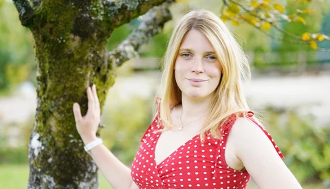 Young woman in red polka dot dress posing with hand on moss-covered tree trunk in garden setting.