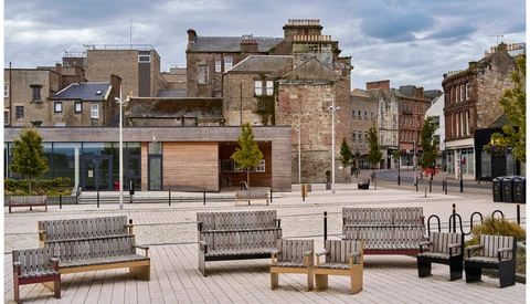 Contemporary public plaza with metal benches and wooden seating arranged on light-colored pavement, facing historic stone buildings.