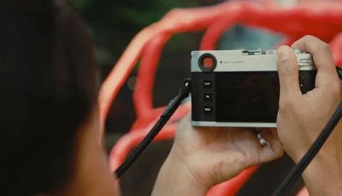 Photographer holding a rangefinder camera with a wrist strap while wearing a red jacket.