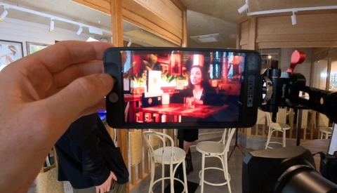 Hand holding smartphone displaying video content in a studio setting with red lighting and seated figures.