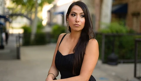 Woman in black sleeveless dress posing outdoors on urban street with blurred background.