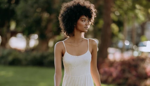 Woman in white camisole dress posing outdoors among trees and grass.