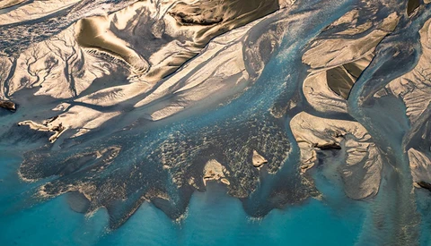 Aerial view of braided glacial rivers flowing through tan and brown terrain into turquoise water.