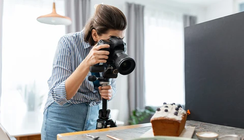 Photographer holding a professional camera with telephoto lens on a tripod while composing a food still life shot.