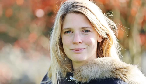 Woman with blonde hair wearing dark jacket with fur collar, photographed outdoors during golden hour.