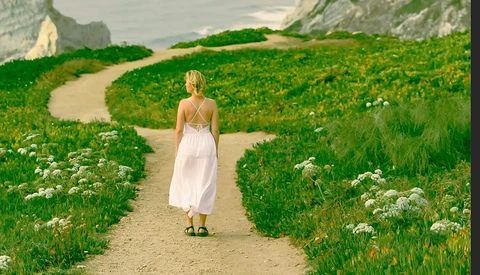 Woman in white dress walking down a dirt path through rolling green fields with white wildflowers.