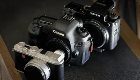 Three digital cameras arranged on a dark surface: a Canon EOS 5D Mark IV, a Fujifilm X-T4, and a compact mirrorless camera in silver.