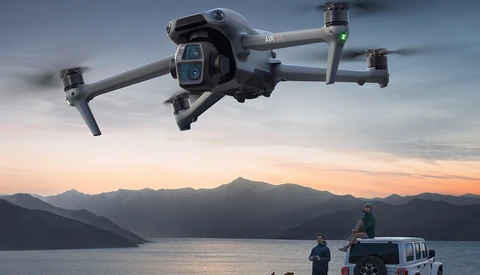 Advanced drone hovering above a vehicle parked on a pier with mountains and water in the background at sunset.