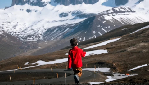 Runner in red jacket jogging on mountain road with snow-capped peaks and patches of snow in background.