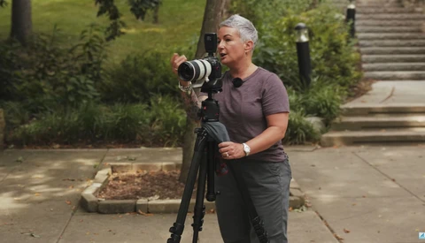 Photographer holding a telephoto lens on a tripod while composing a shot outdoors.