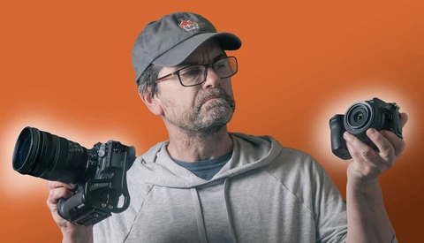 Videographer holding cinema camera in left hand and compact camera in right hand against orange background.