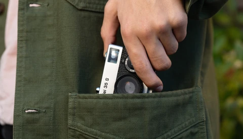Hand holding a compact rangefinder camera with white body and black lens protruding from olive cargo pants pocket.