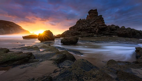Rocky coastline at sunset with golden light between layered stone formations and smooth water in foreground.