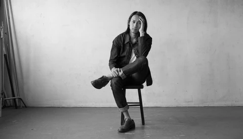 Person sitting on a wooden chair in a minimalist studio setting, resting chin on hand in a contemplative pose.