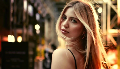 Woman with long reddish-blonde hair looking over shoulder in urban evening setting with warm city lights.