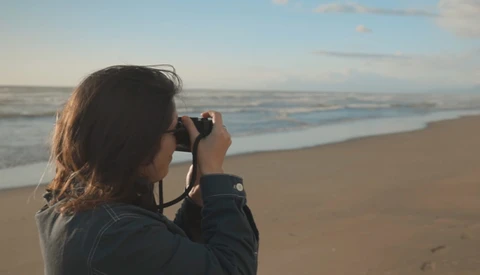 Woman photographing the ocean from a beach during golden hour.