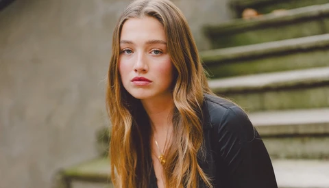 Young woman with long reddish-brown hair wearing black jacket, posed near moss-covered concrete steps.
