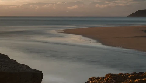 Serene coastal landscape at dusk with smooth water, sandy beach, and rocky outcrops.