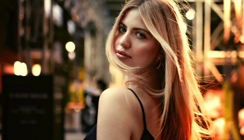 Woman with long reddish-blonde hair photographed outdoors at night with warm city lights in background.